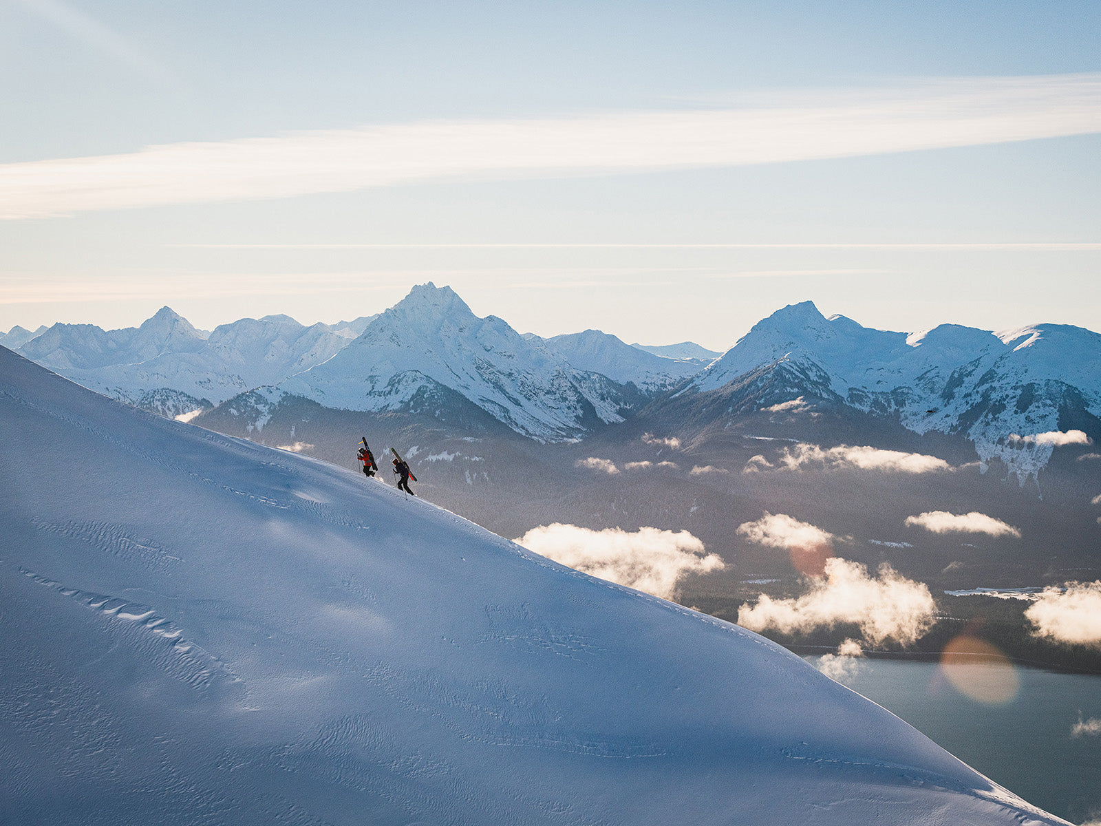 Ellen Bradley and Connor Ryan bootpack up a ridge near the ski area, with the peaks of Lingít Aaní (Tlingit land) towering in the distance.